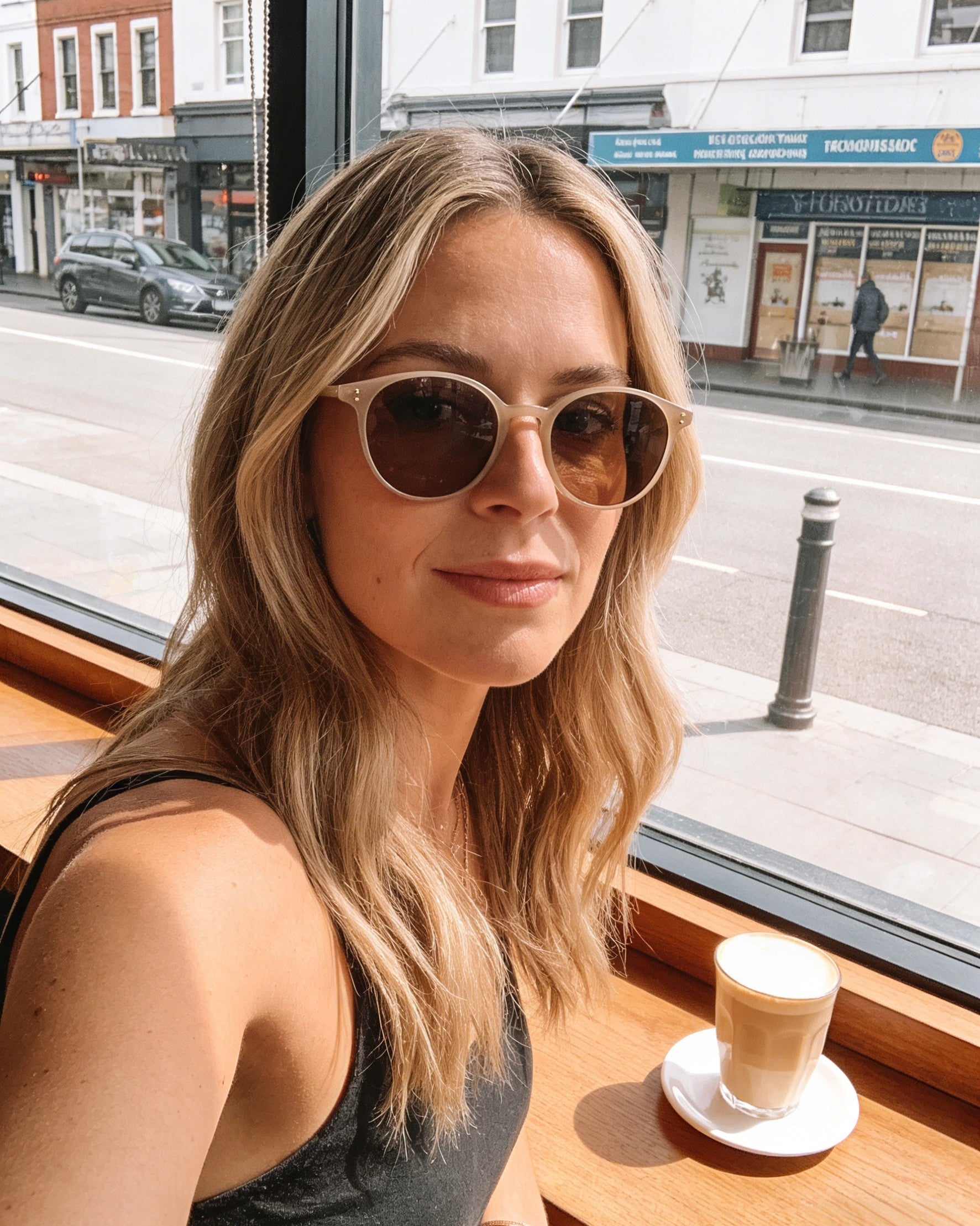 Woman wearing sunglasses sitting at a table with a coffee cup in a cafe. Classic round unisex beige color sunglasses | polarised lenses, 100% UV protection, stylish trendy fashion